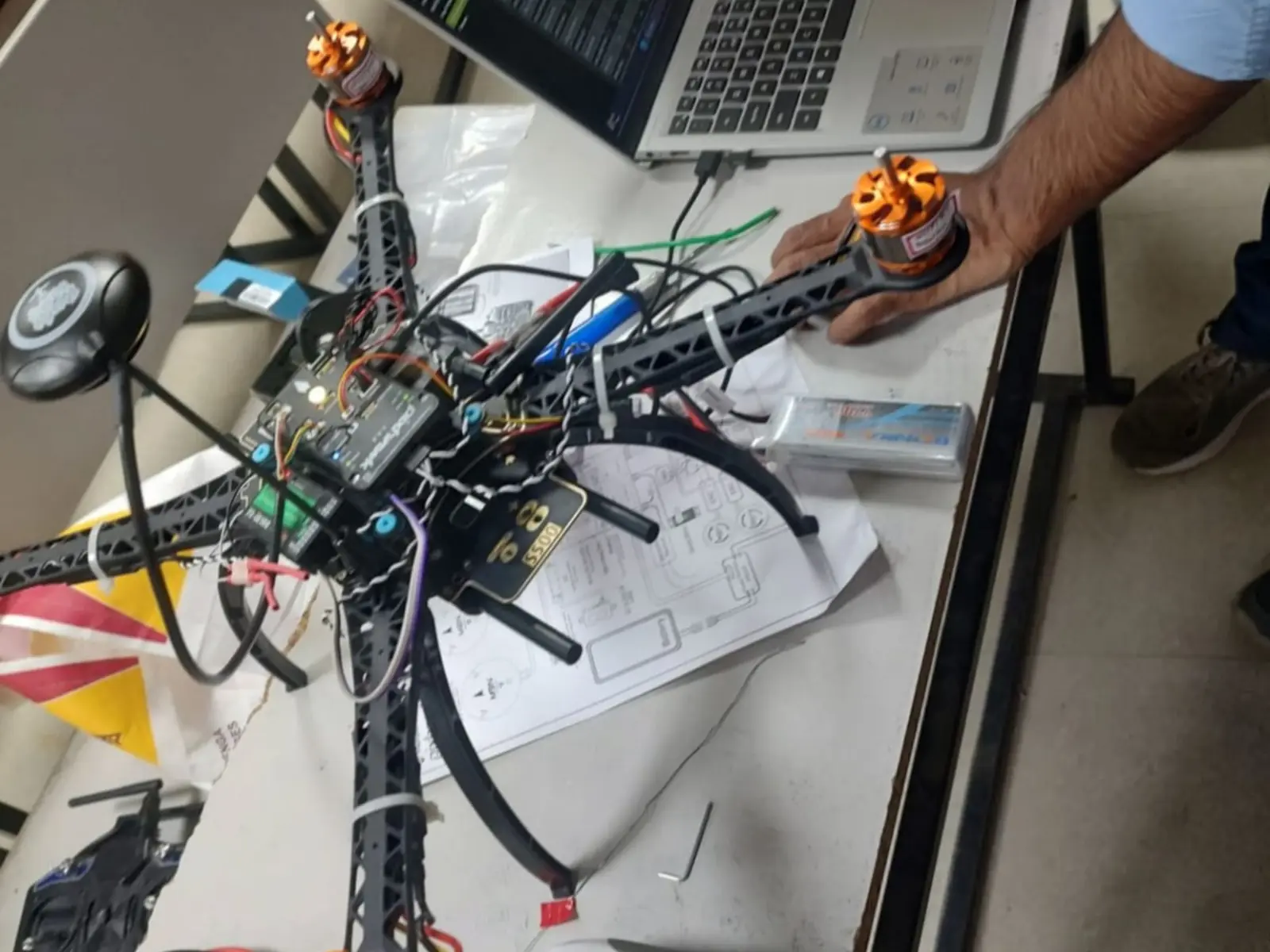 A quadcopter under assembly beside its controller, laptop and engineering diagrams.